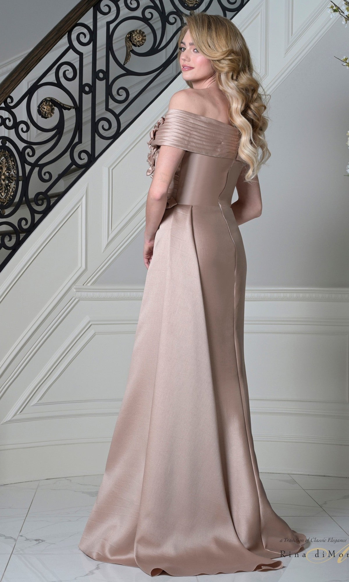 A woman with long blonde hair wears the Long Formal Dress: Rina Di Montella RD3528—an off-shoulder, floor-length beige satin gown with 3D floral appliques—standing on a white marble floor before an ornate staircase.